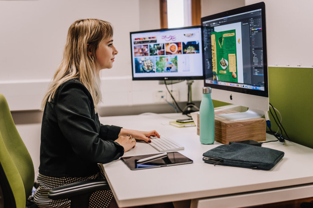 Side profile of a female wearing a black jacket sat at her desk working from two computer screens